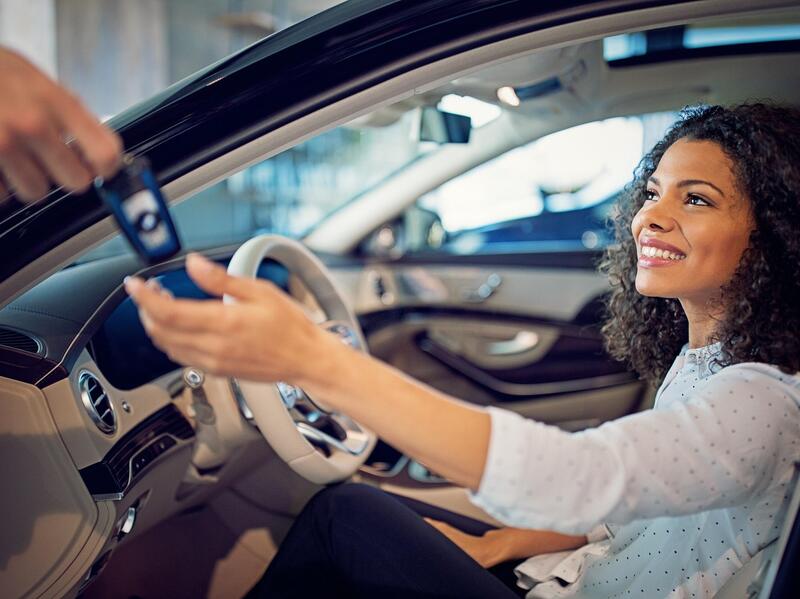 A woman sitting in a car with an open door smiling and reaching out to a hand giving her a car key