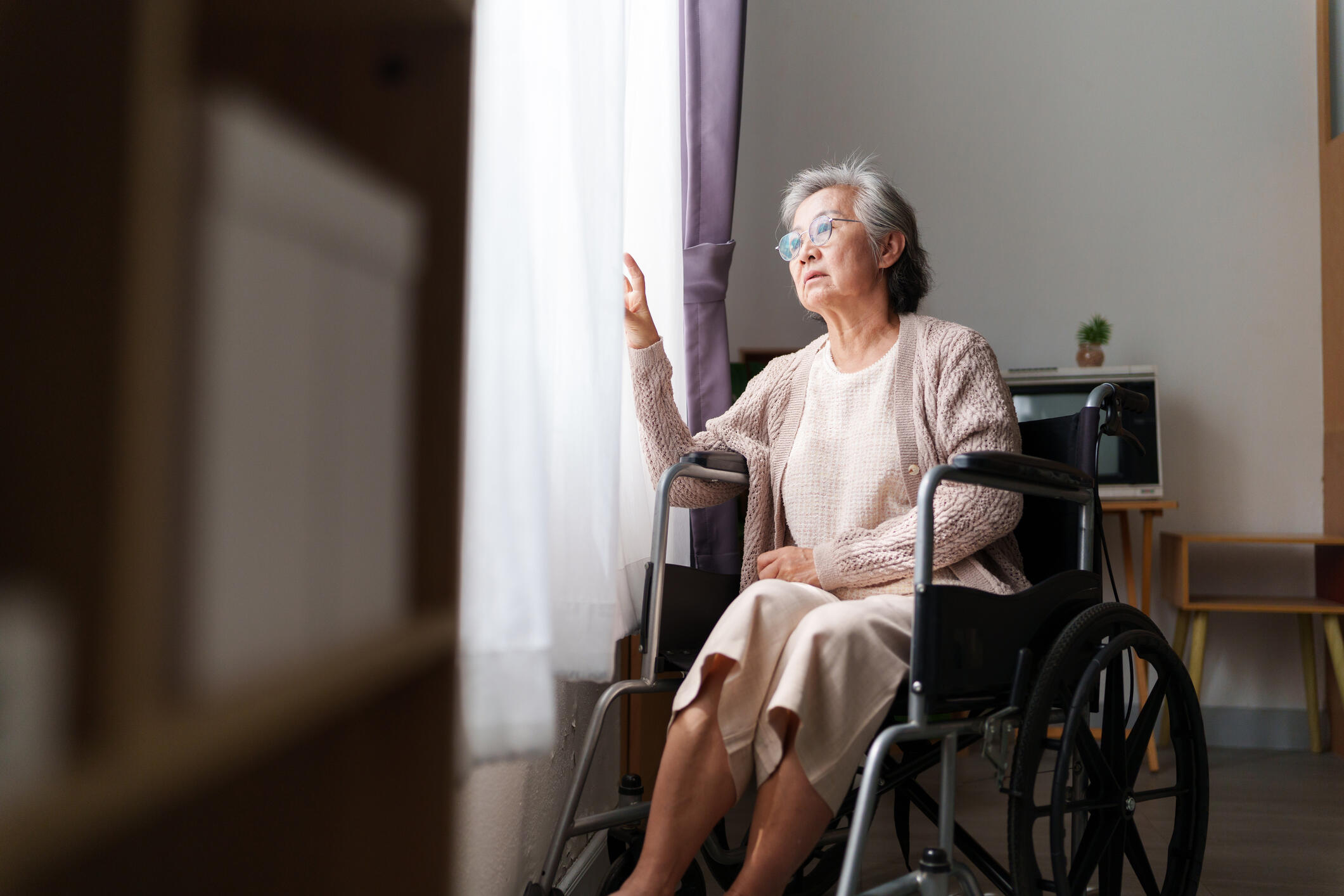 A photo of an older woman looking out a window while sitting in a wheel chair. 