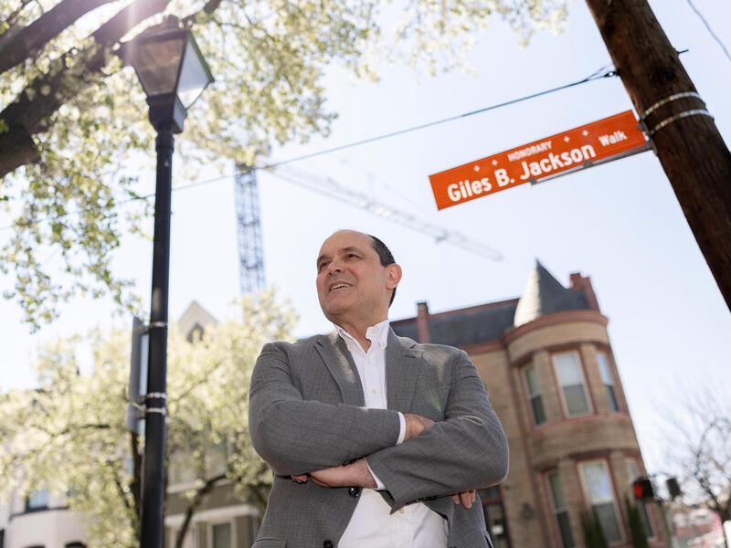 A photo of a man standing at a street corner with his arms crossed against his chest. 