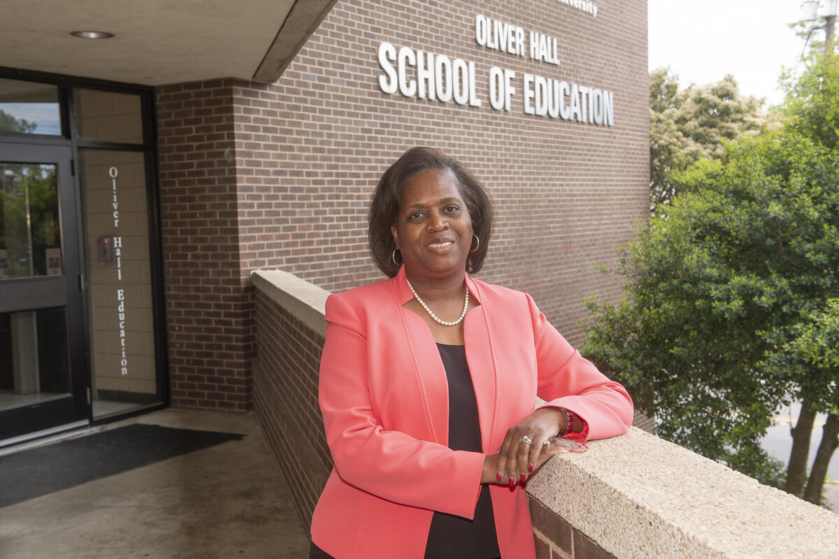 A photo fo a woman fromt he waist up leaning her right arm against a brick wall. Behind her is a building with white text on the side that reads \"OLIVER HALL SCHOOL OF EDUCATION\" 