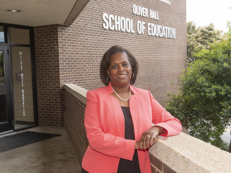 A photo fo a woman fromt he waist up leaning her right arm against a brick wall. Behind her is a building with white text on the side that reads \"OLIVER HALL SCHOOL OF EDUCATION\" 