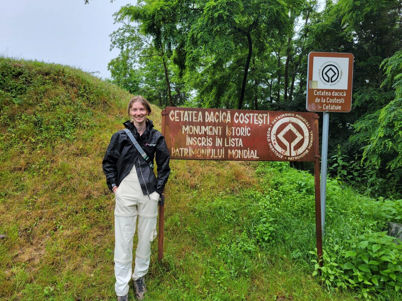 A photo of a woman standing on a grassy hill next to two brown signs. Both of the signs are written in a foreign language. 