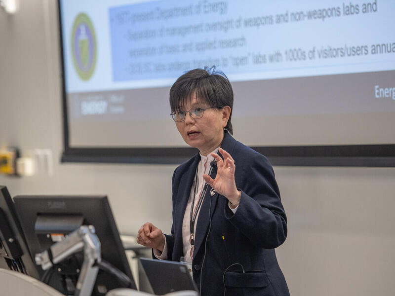 A photo of a woman standing behind a podium with a computer screen on it. She is speaking and has her right hand raised in front of her. 