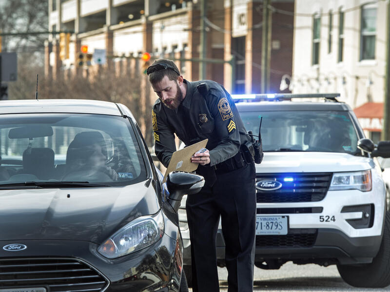 A police officer standing next to a car talking to the driver. The officer is holding a clip board and showing it to the driver. There is a police suv behind them with the lights on 