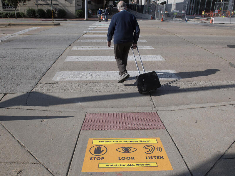 A pedestrian pulling a rolling briefcase begins to cross a street at a crosswalk. A sign placed on the sidewalk states \"Hands Up & Phones Down,\" symbols indicating pedestrians to \"STOP\" \"LOOK\" and \"LISTEN,\" and \"Watch for ALL Wheels.\"