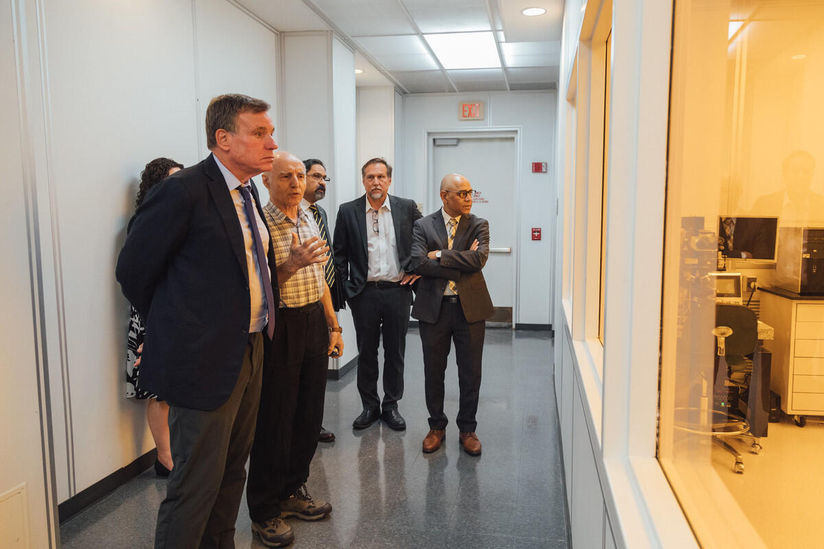 A photo of several people standing in the hallway of a building, looking into windows that show another room. 