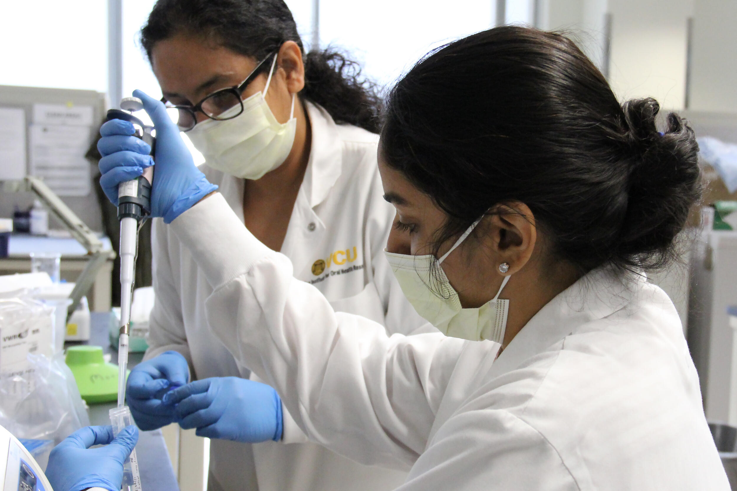 Two women wearing lab coats, facemasks, and rubber glovers working