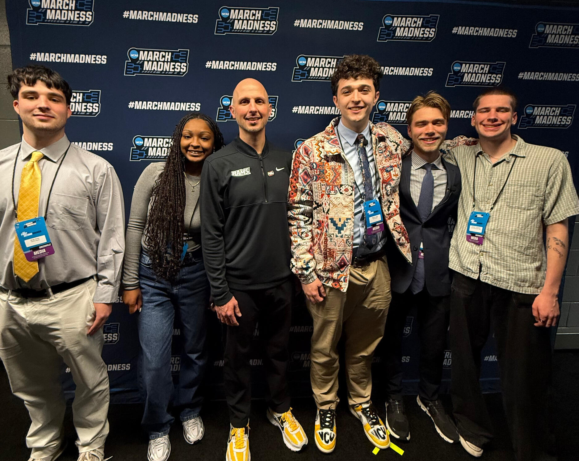 A group of six people pose in front of a blue \"March Madness\" backdrop.
