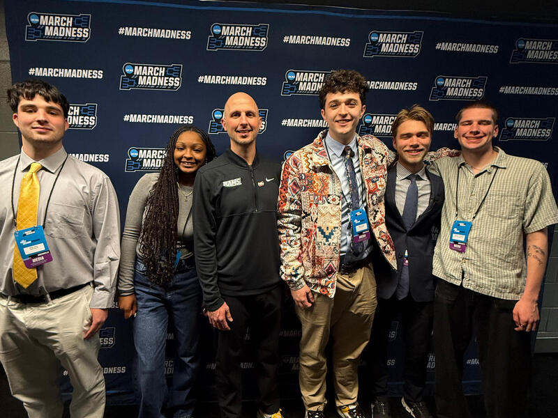 A group of six people pose in front of a blue \"March Madness\" backdrop.