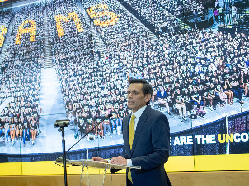 Photo of a man in a suit with a yellow tie speaking at a lectern in front of an image of a large crowd of sitting people.