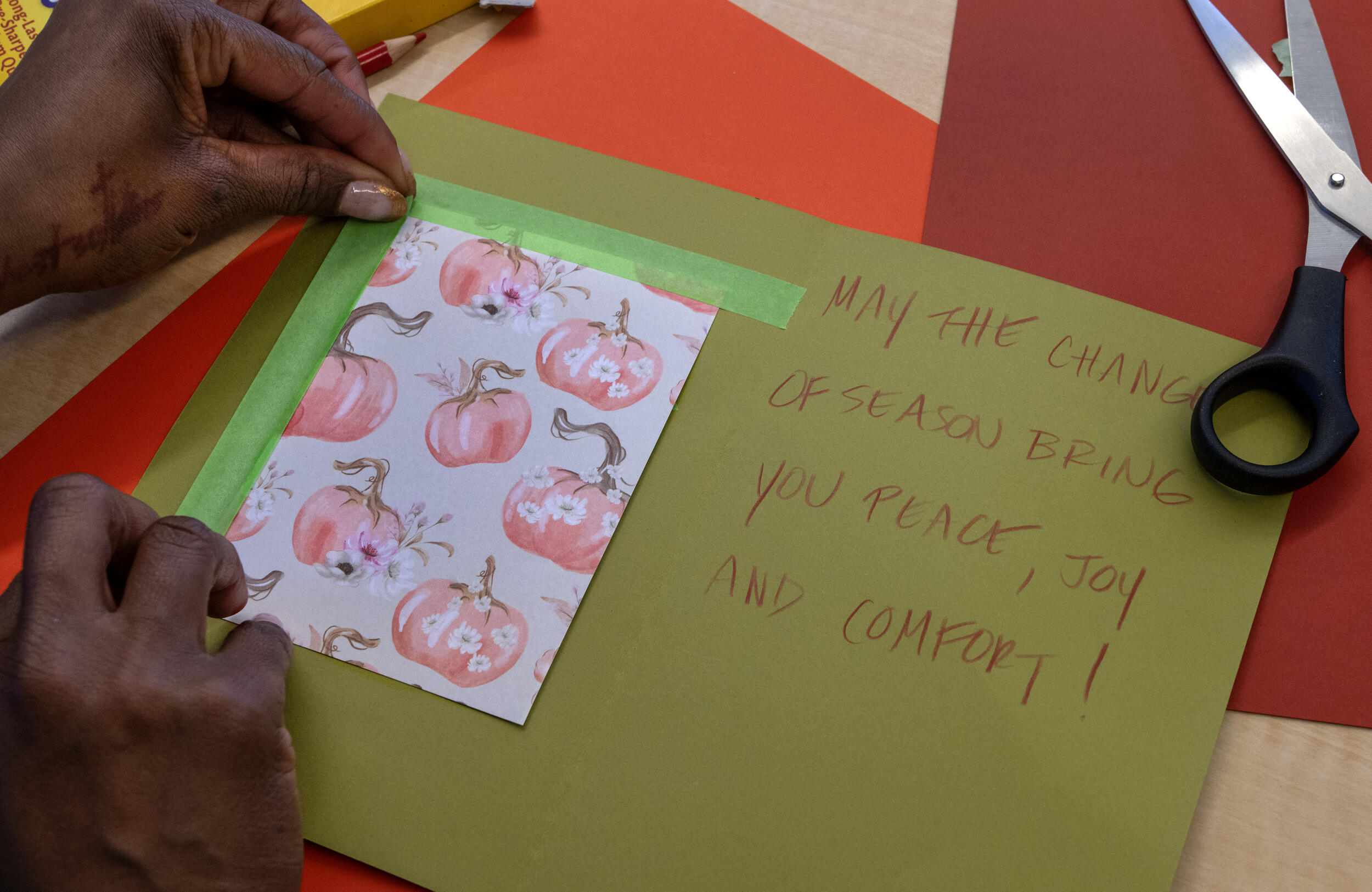 A photo of a card with two hands taping down a piece of paper with illustrations of pumpkins on it. The right of the card says \"MAY THE CHANGE OF SEASON BRING YOU PEACE, JOY AND COMFORT!\"