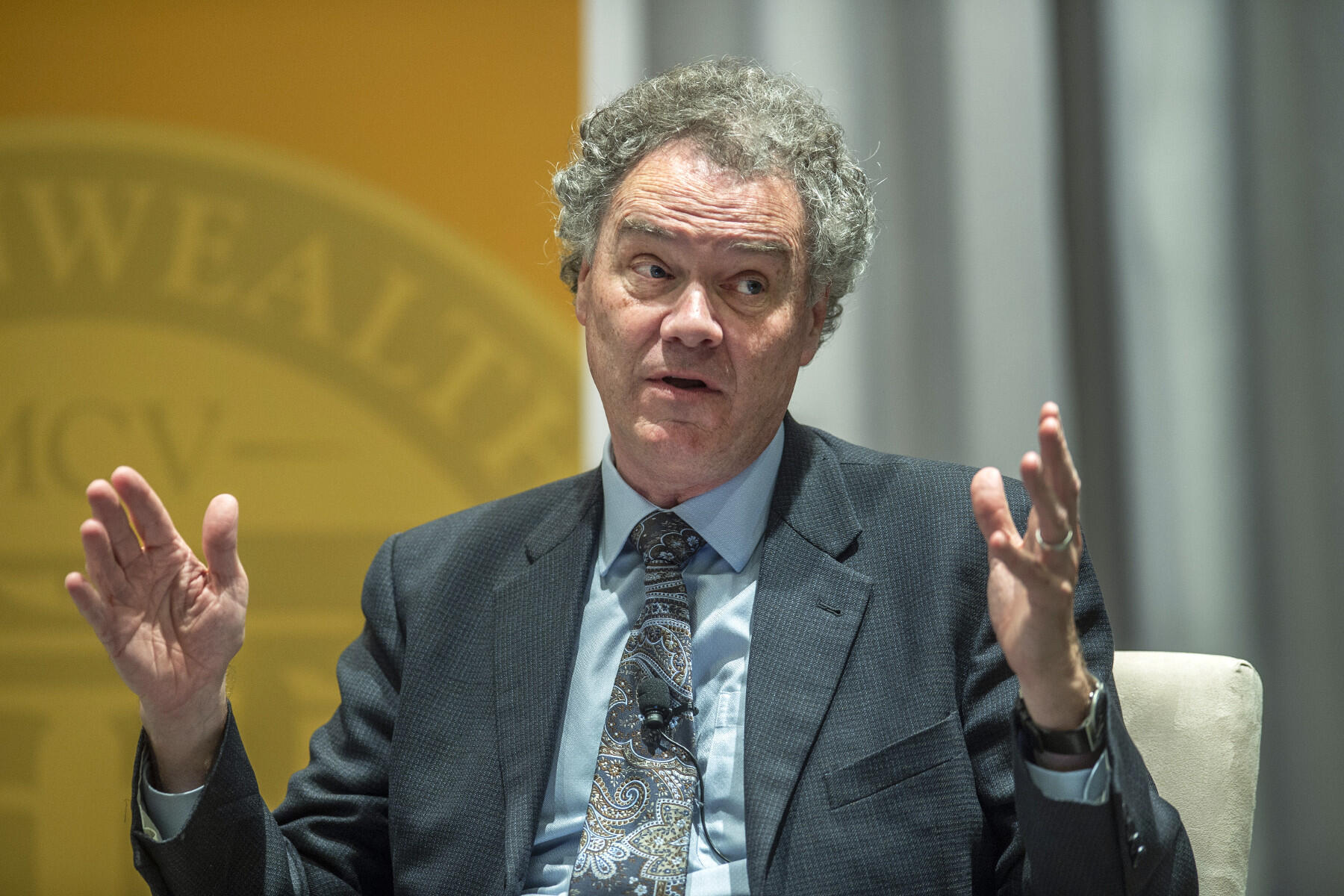 A man in a suit holds up his hands while speaking.