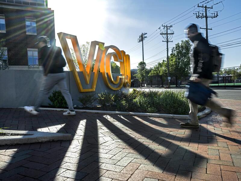 A photo of wo people walking on a brick sidewalk past a sign made up of giant yellow letters that spell out \"VCU.\" 