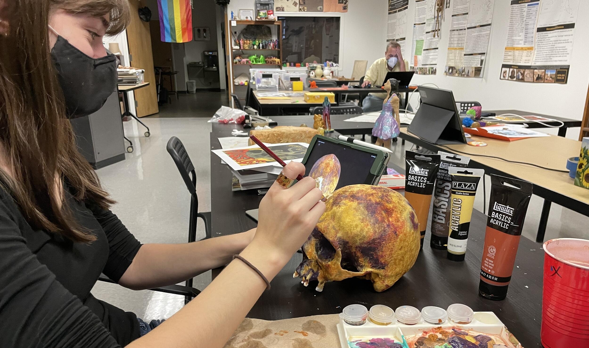 A woman wearing a face mask painting a skull at a table 