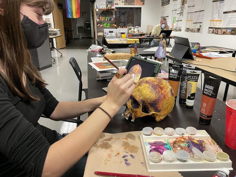 A woman wearing a face mask painting a skull at a table 