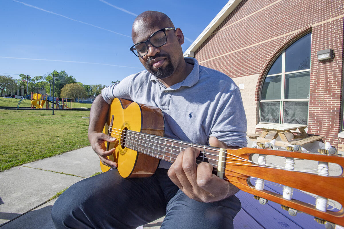 A man playing an acoustic guitar while sitting on a bench. Behind him on his right is a brick building and on the left is a playround. 