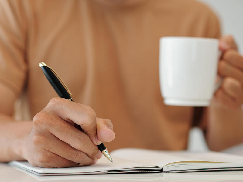 A photo of a man sitting at a table writing in a notebook while holding a cup of coffee. 