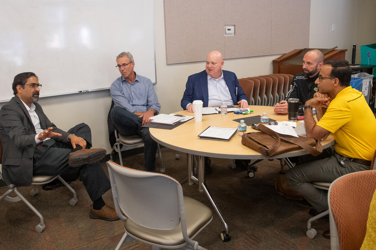 A photo of four men sitting around a circular table, and one man sitting to the left to the table. The four men at the table are looking at the man sitting to the left. The man sitting to the left is speaking. 
