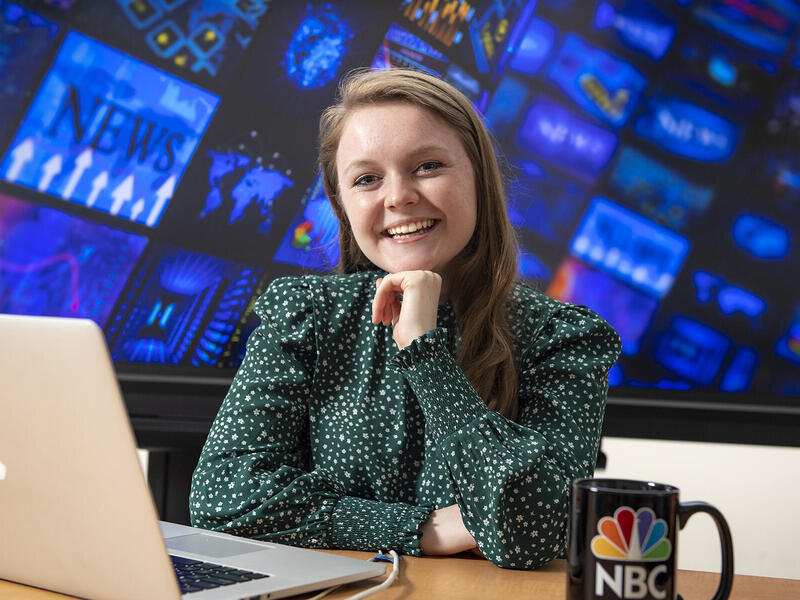 Allison Martin sitting at a desk with a laptop in front of her and a coffee mug that says \"NBC.\" 