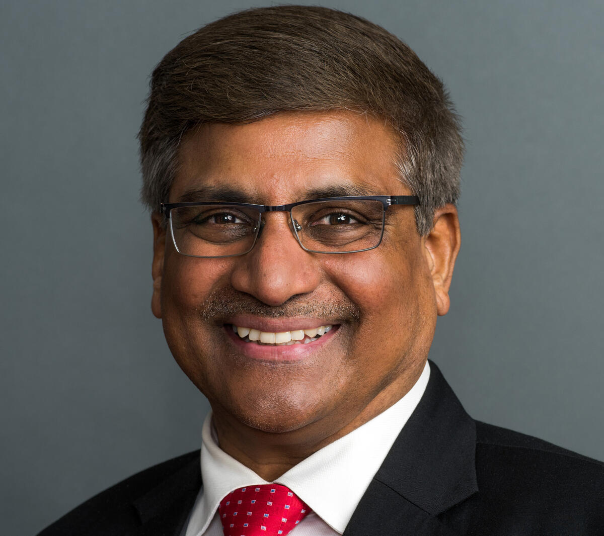 A photo of a man wearning a black suit jacket, a white undershirt, a red tie, and glasses. 
