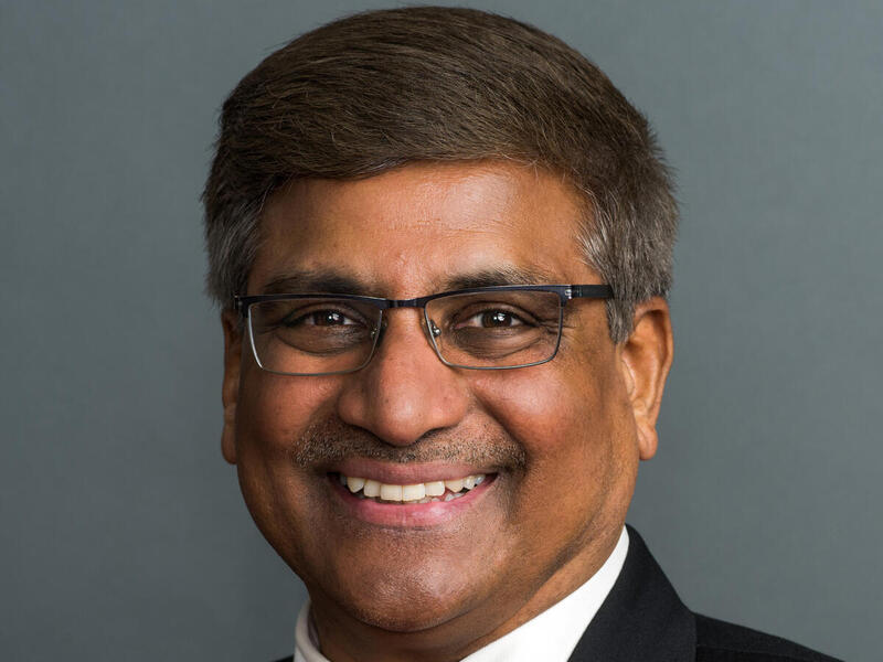 A photo of a man wearning a black suit jacket, a white undershirt, a red tie, and glasses. 