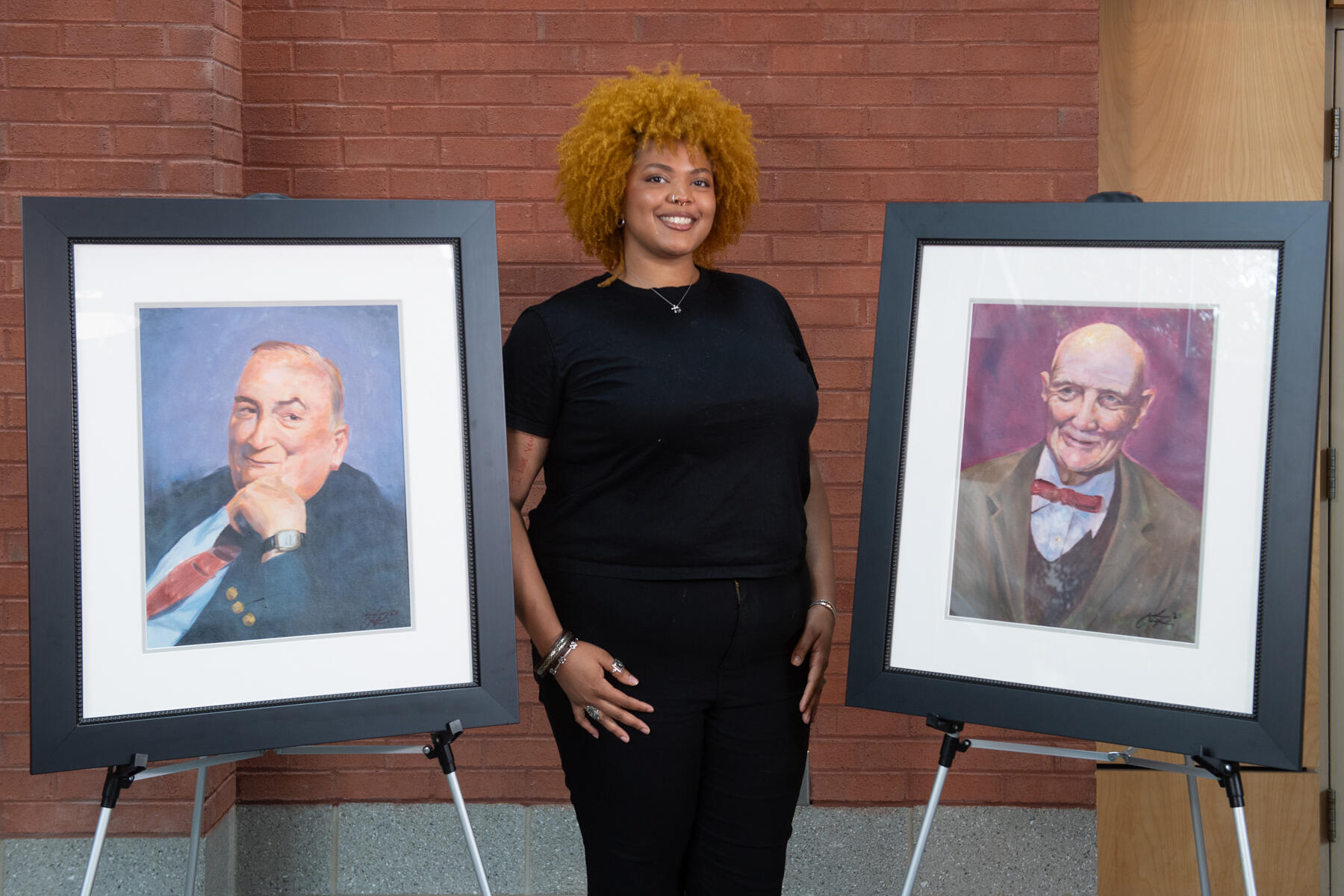 A woman smiles between two framed portraits of men.