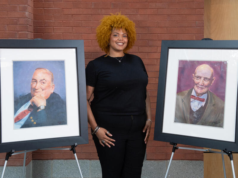 A woman smiles between two framed portraits of men.
