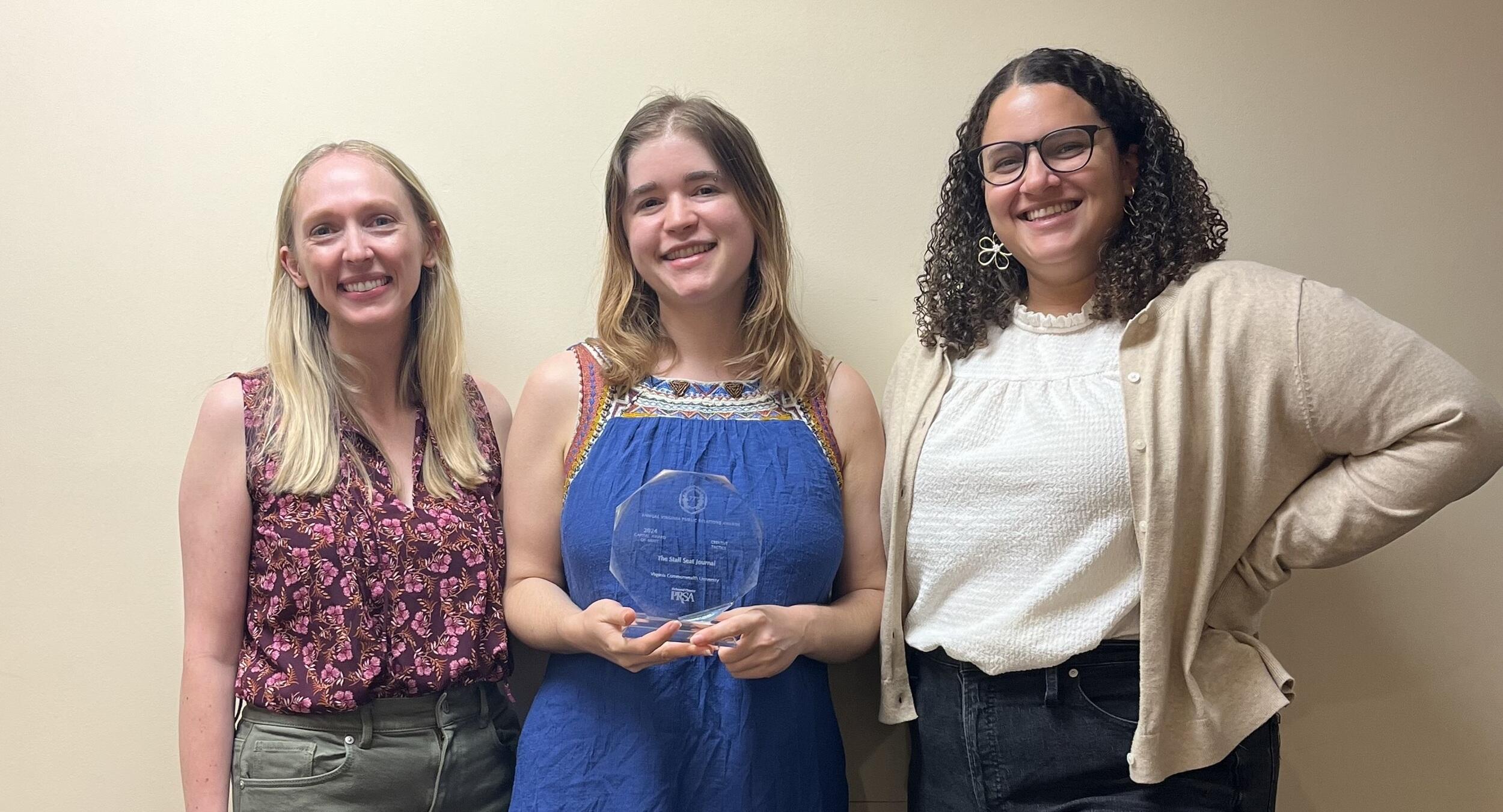 A photo of three people standing. The person in the middle is holding a glass trophy