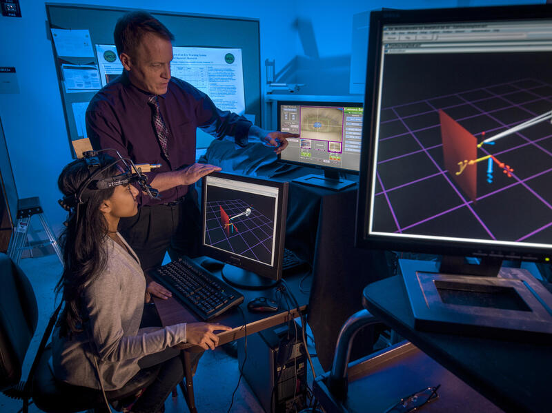 A woman sitting in front of a computer with a headband device on her head. Next to her is a man pointing at another computer screen. 