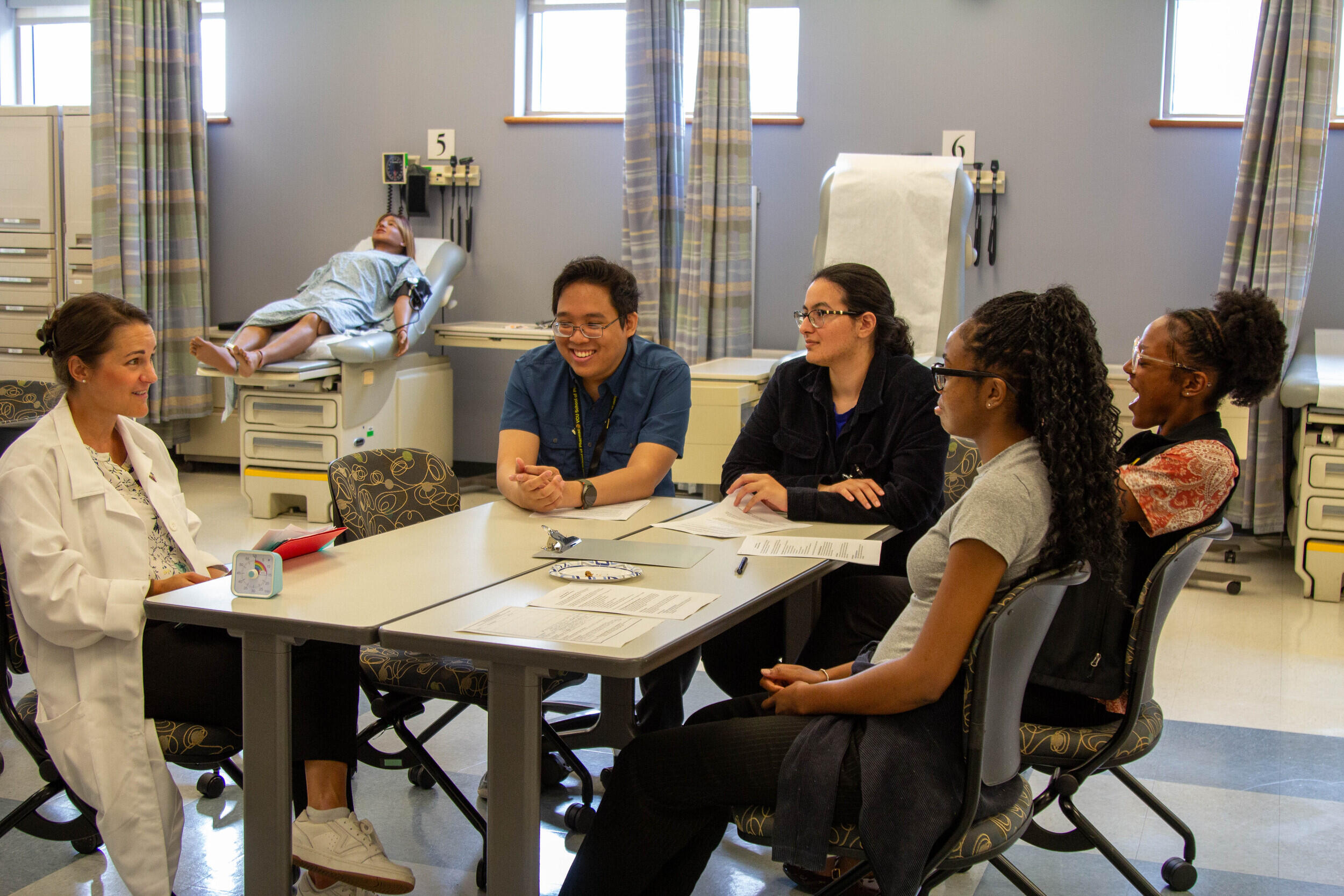 A photo of five people sitting around a table. There is a dummy laying on a hospital bed behind them. 