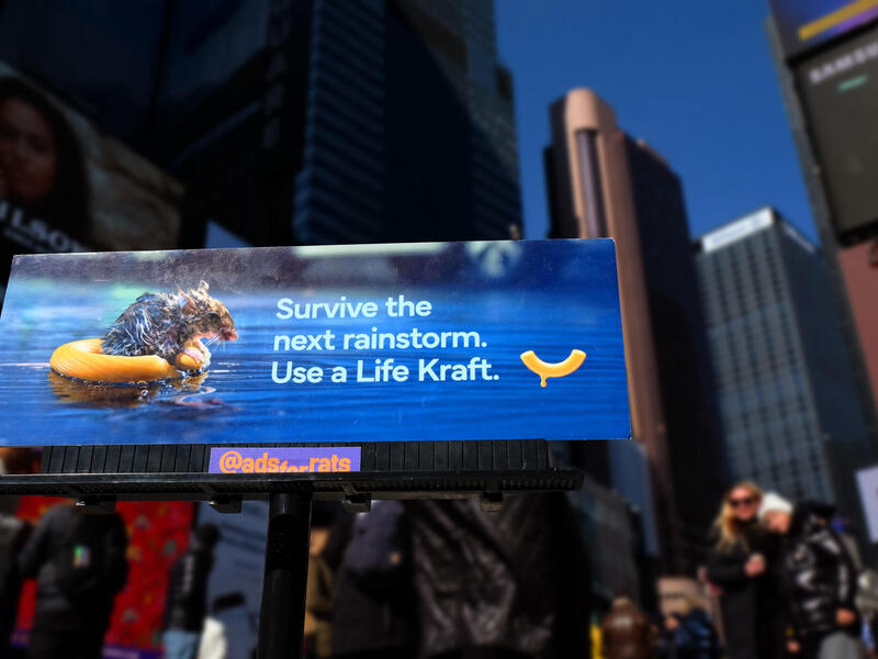 A photo of a small billboard with a photo of a rat using two macaroni noodles as a flotation device in water. White text reads \"Survive the next rainstorm. Use a Life Kraft.\" 