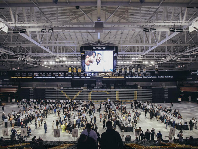 A phot of the basket ball court in the Siegel Center's basketball court floor full of tables with posters on them. The room is full of people around the booths, and a few people are sitting in the bleachers. 