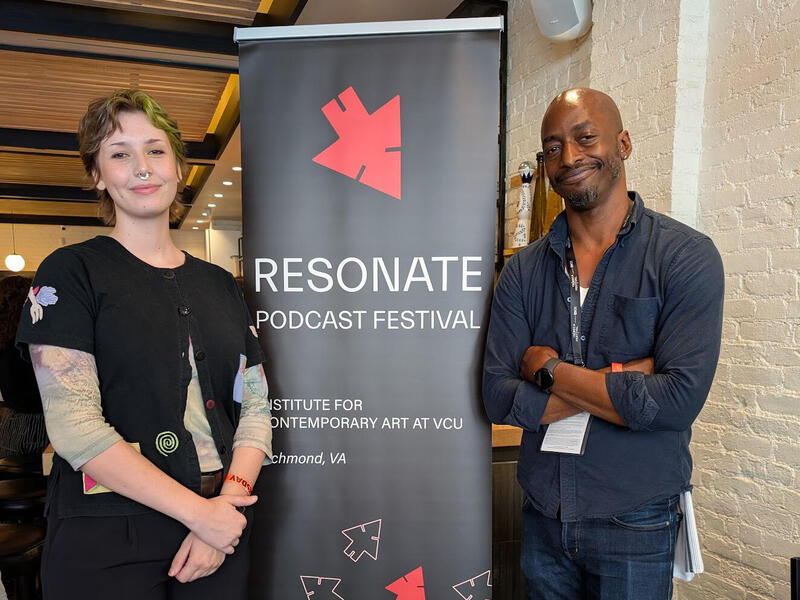 A photo of two people standing on either side of a black banner with white text that reads \"RESONATE\" \"PODCAST FESTIVA\" \"INSTITUT FOR CONTEM PORARY ART AT VCU\" \"Richmond, VA\"