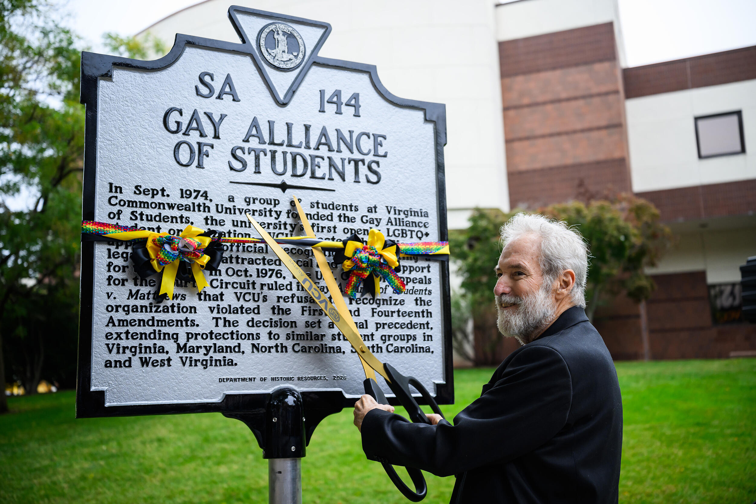 A photo of a man cutting yellow, black, and rainbow ribbons wrapped around a sign with a large pair of scissors. The top of the plaque says \"SA\" \"144\" \"GAY ALLIANCE OF STUDENTS.\"