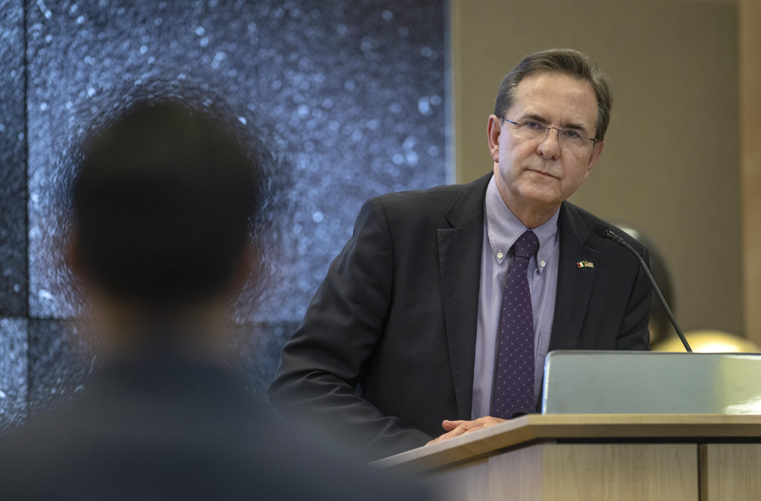A photo of a man standing at a podium looking at a person in front of him. 