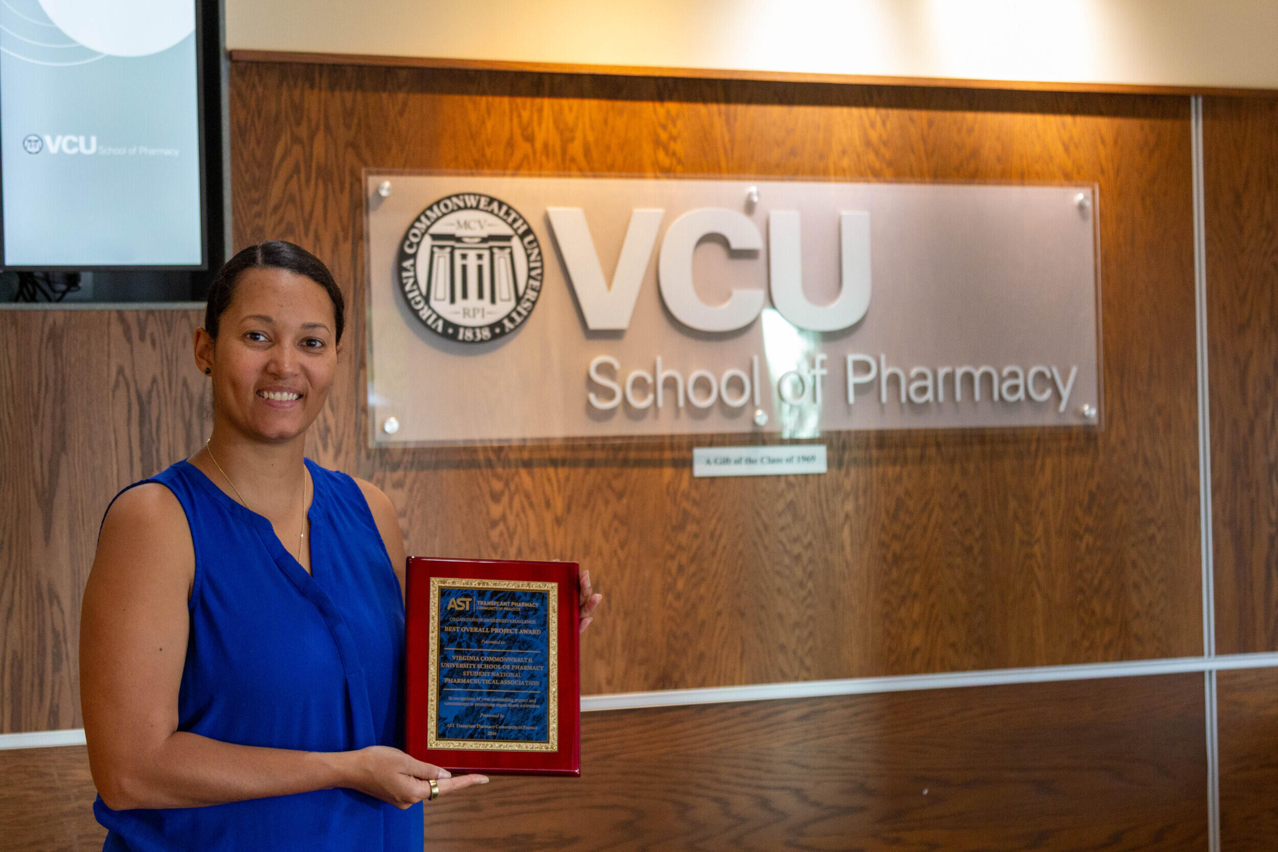 A photo of a woman from the waist up. She is holding an award plaque. The woman is standing to the left of a sign that says \"VCU School of Pharmacy.\" 