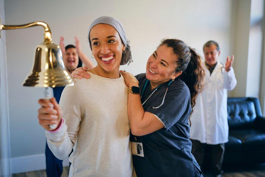 A photo of a woman smiling and ringing a bell. Next to her a nurse is holding onto her sholder. Behind them another man and woman are clapping. 