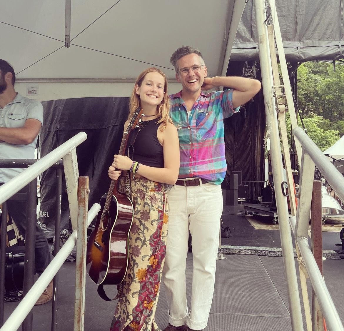 A photo of a woman standing on the left holding a guitar and a man standing to the right of her. Both people are smiling. 