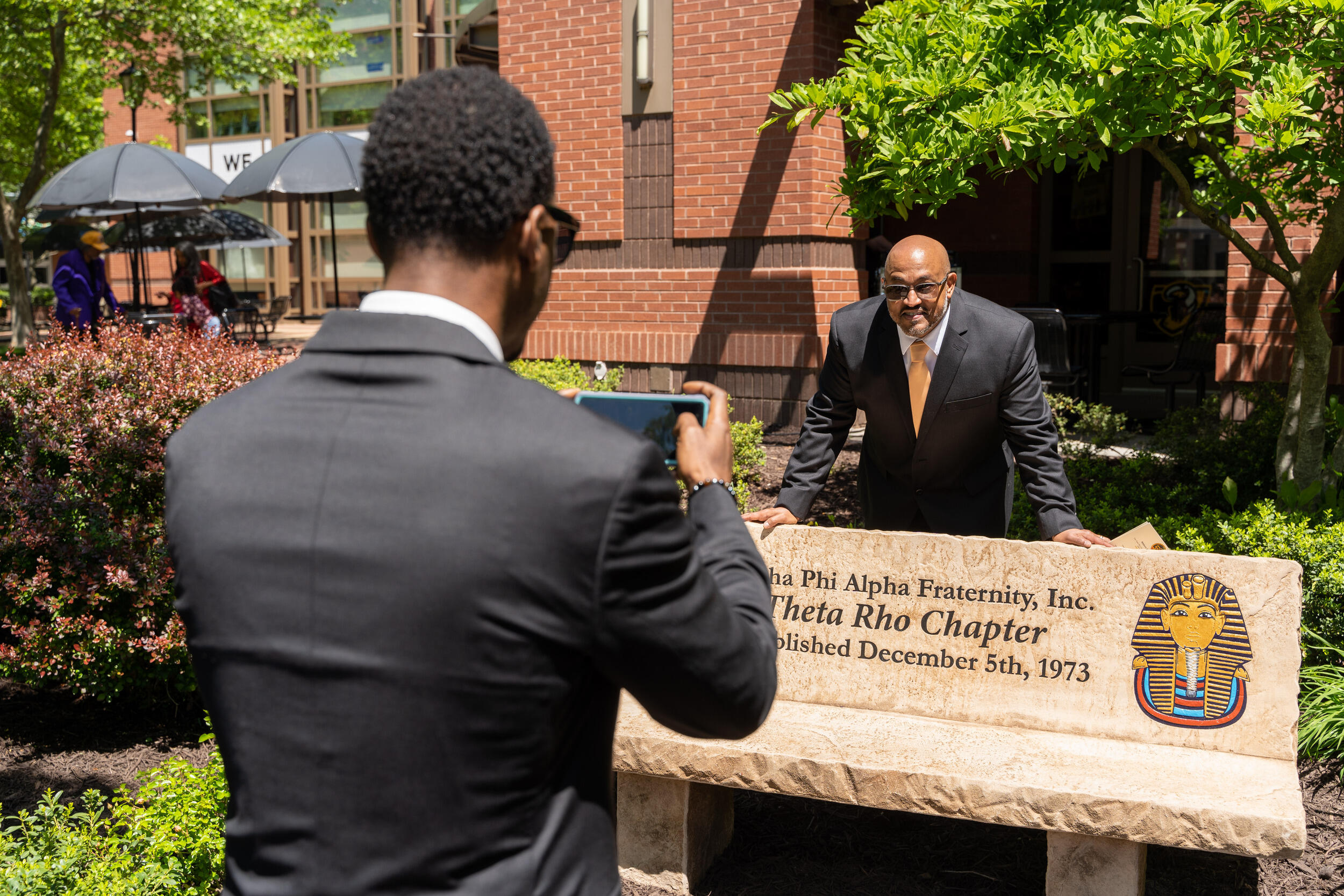 A man posing behind a stone bench while a man in front of him takes a picture with a smart phone. 