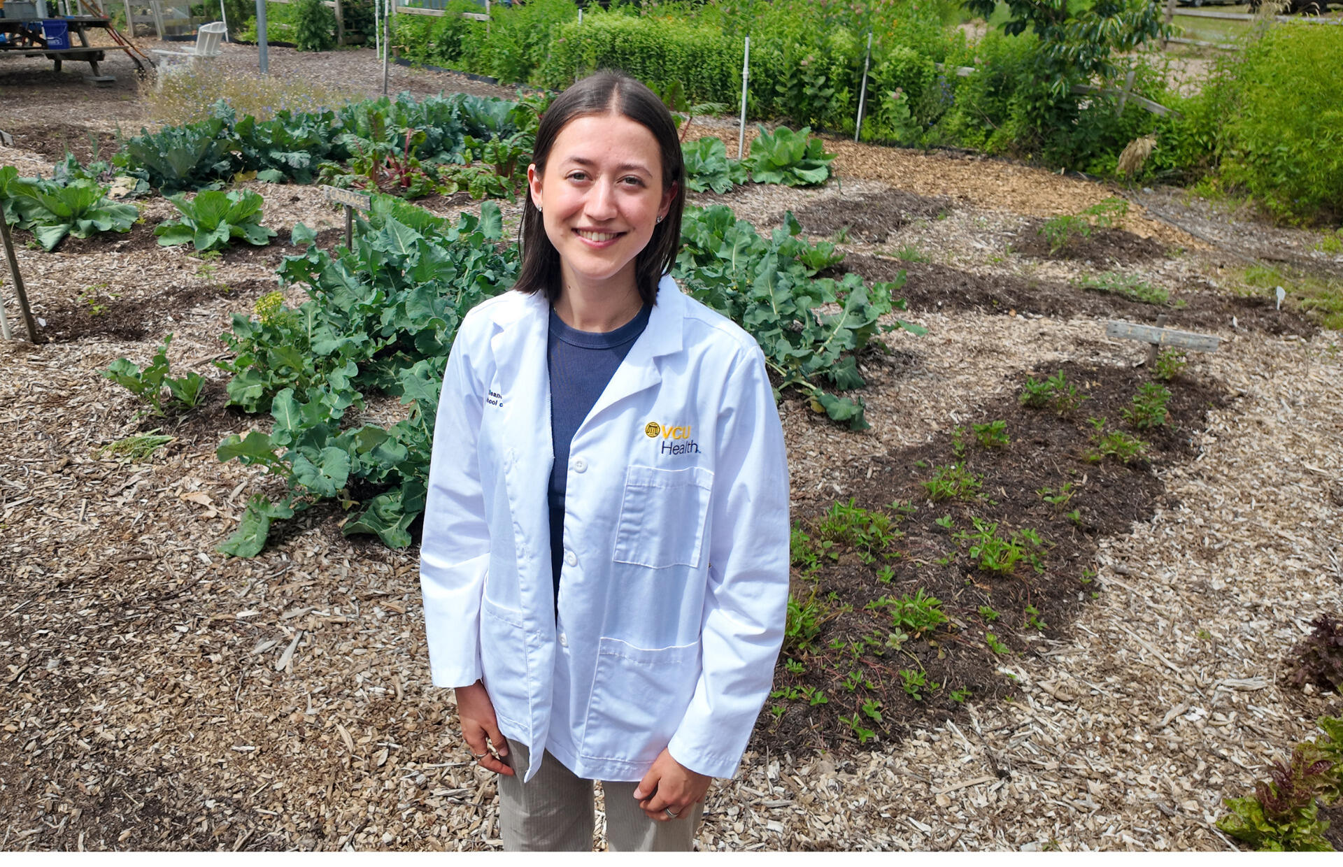 A photo of a woman wearing a white medical coat that says \"VCU Health\" standing in a vegtable garden. 