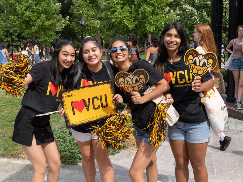 A photo of four women stading next to eachother holding pom poms, a sign that says \"I heart VCU,\" and paper rodney the ram masks. 