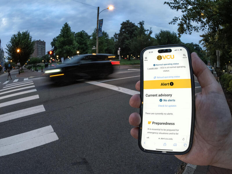 A photo of a hand holding a smart phone. The screen shows the VCU Alert page on it. Behind the hand is a street with a blurry car going by. 
