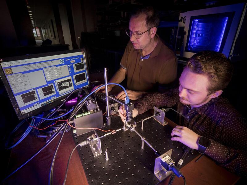 Two men standing next to each other in front of a table. The man on the left is looking at a computer screen and the man on the right is looking at and touching a structure made of metal with wires coming out of it. 