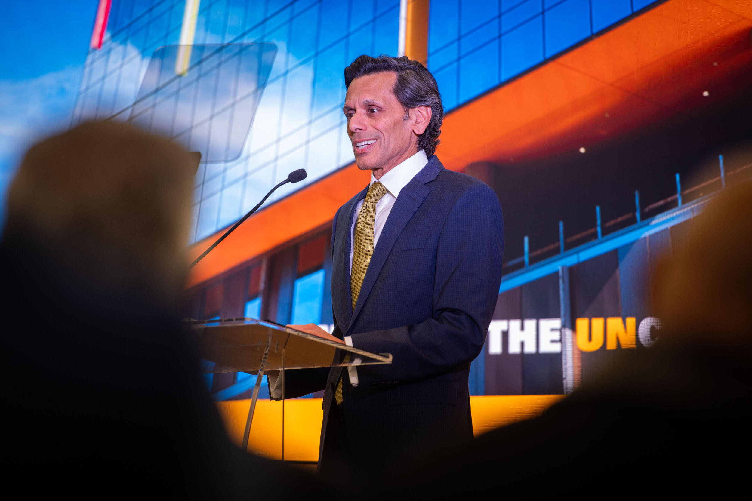 A man in a coat and tie speaks into a microphone at a lectern with the image of a building behind him.