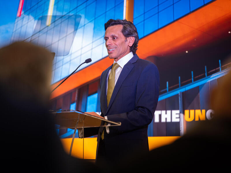 A man in a coat and tie speaks into a microphone at a lectern with the image of a building behind him.