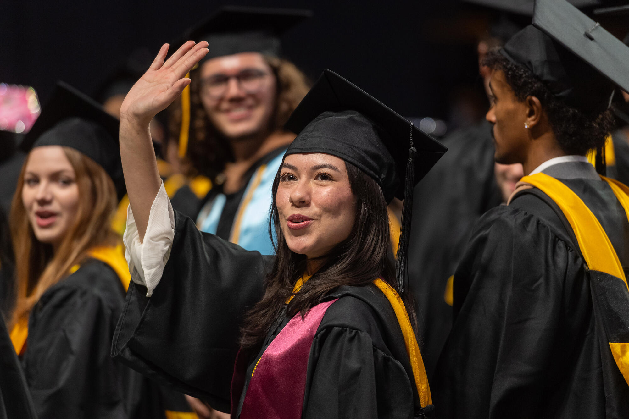 A graduate in a cap and gown waves. Other graduates in cap and gown are in the background.