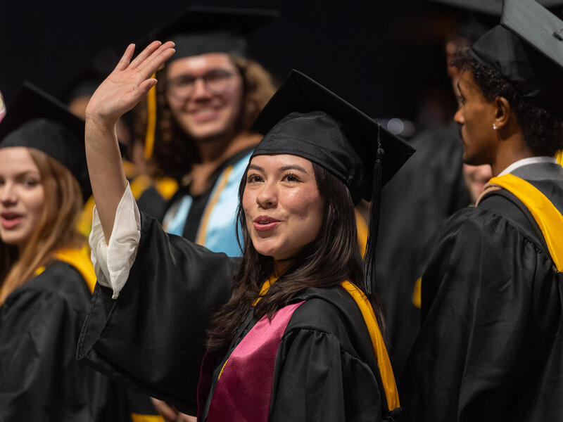 A graduate in a cap and gown waves. Other graduates in cap and gown are in the background.