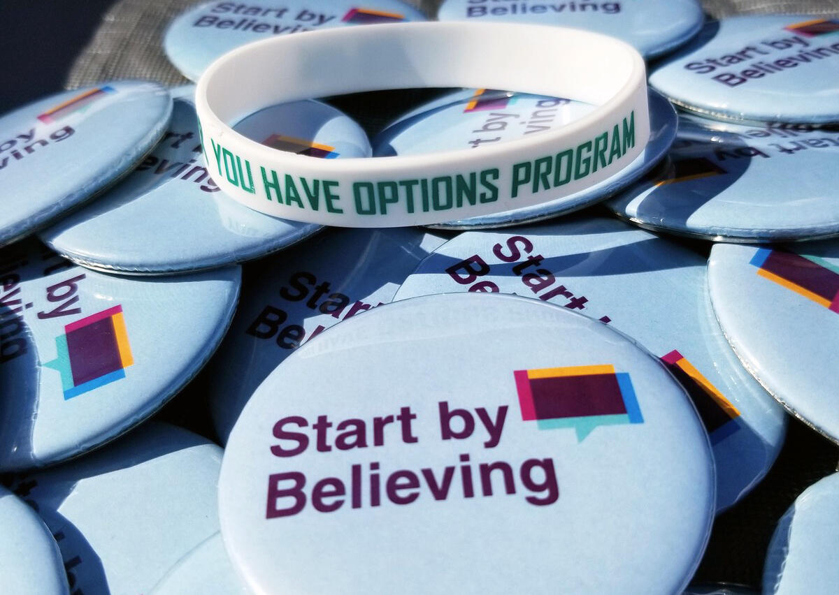 A photo of a plastic white bracelet sitting on a pile of pins. The bracelet says \"YOU HAVE OPTIONS\" in green letter. The circular pins the bracelet is sitting on say \"Start by Believing.\" 