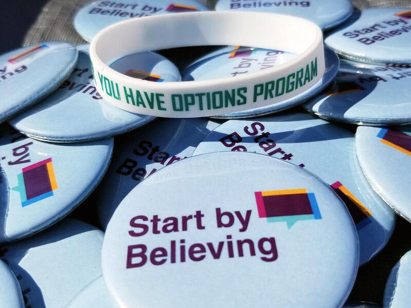A photo of a plastic white bracelet sitting on a pile of pins. The bracelet says \"YOU HAVE OPTIONS\" in green letter. The circular pins the bracelet is sitting on say \"Start by Believing.\" 