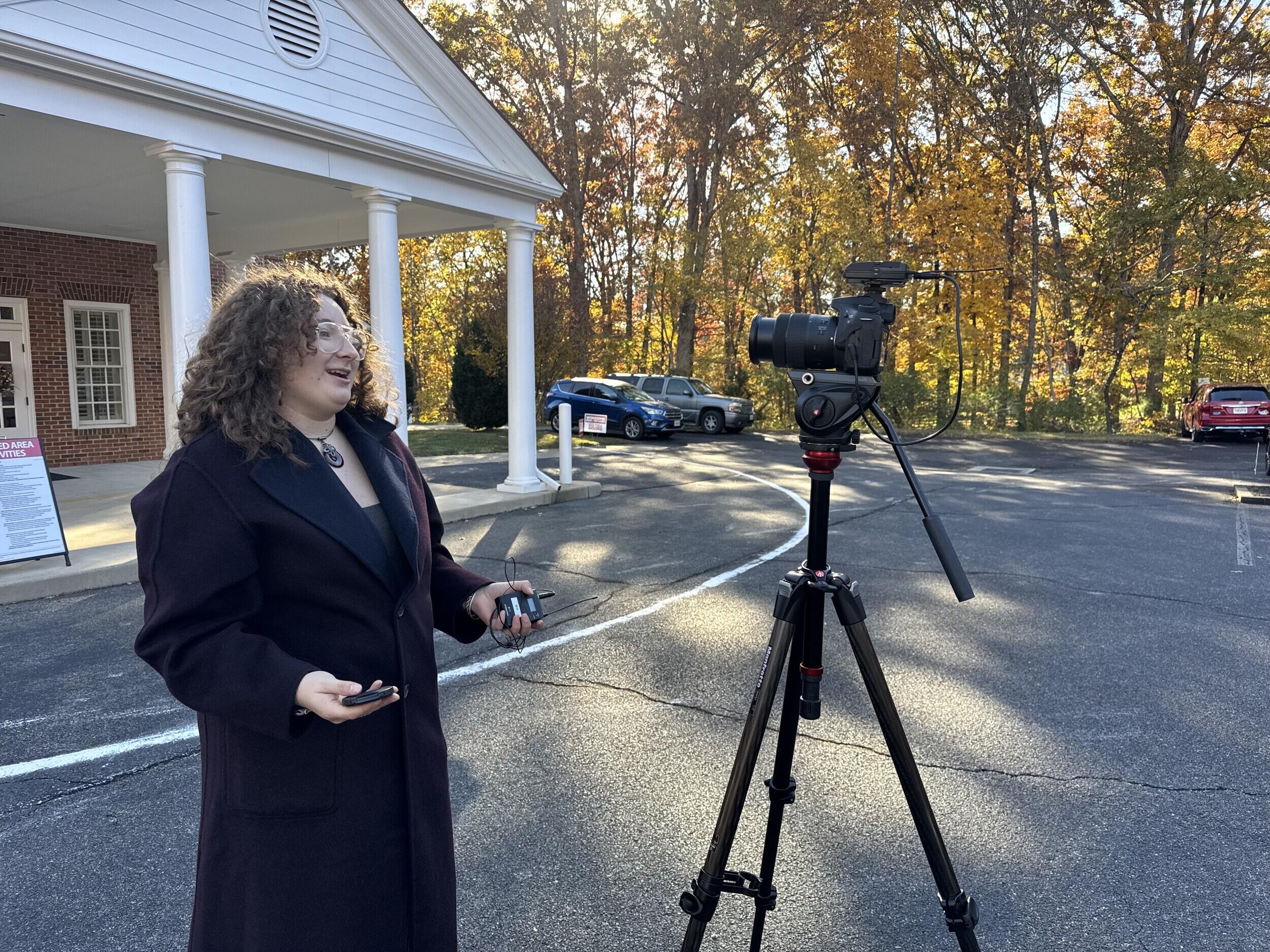 A photo of a woman standing in front of a camera and speaking. 